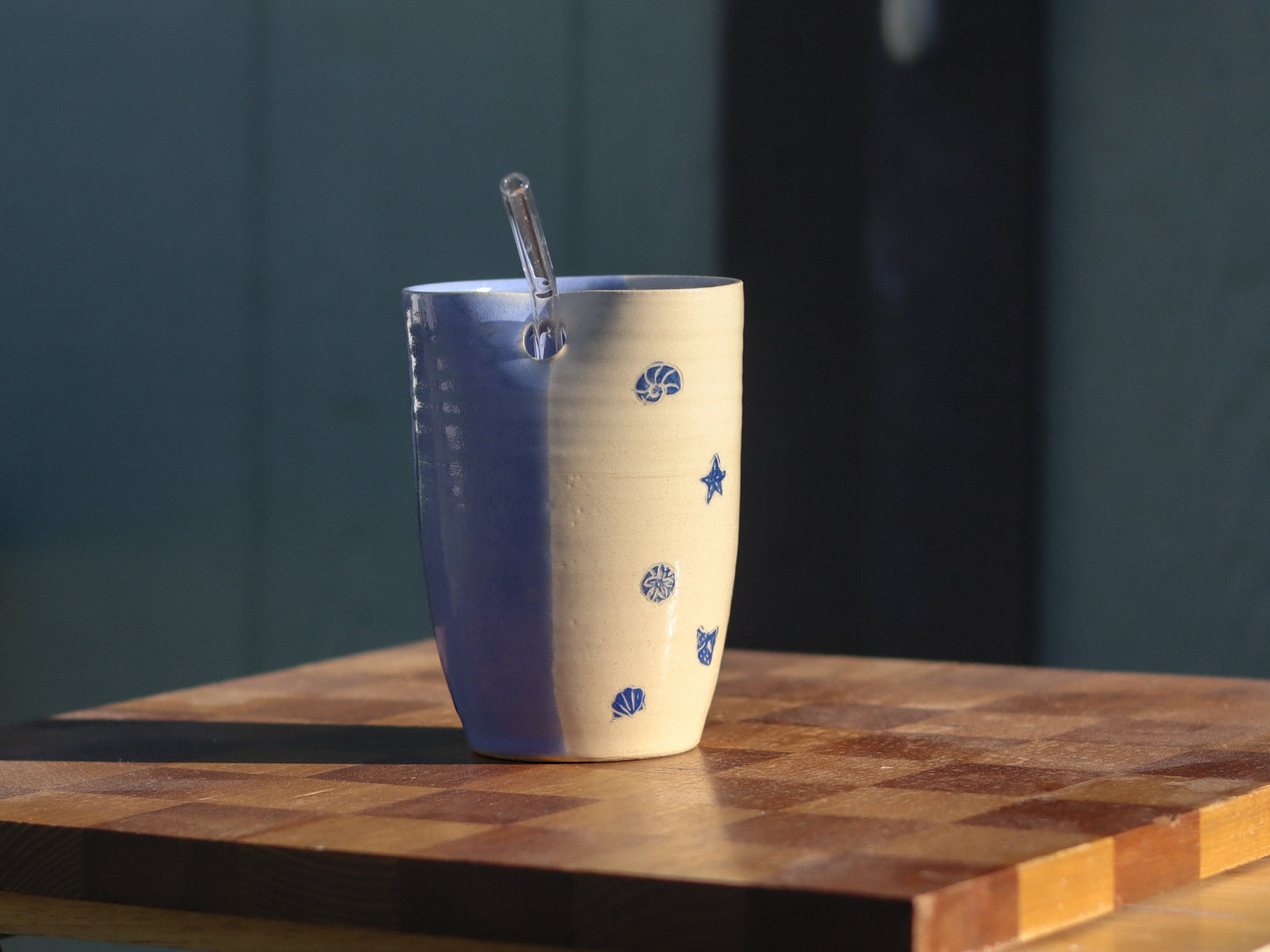 A handmade ceramic straw tumbler with a blue and cream color scheme, decorated with blue seashell and star motifs, sitting on a wooden cutting board with a glass straw inside.