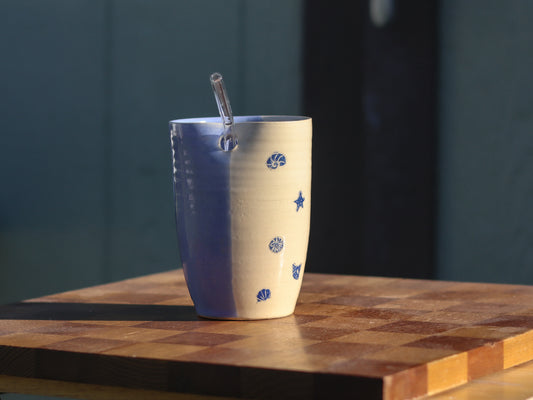 A handmade ceramic straw tumbler with a blue and cream color scheme, decorated with blue seashell and star motifs, sitting on a wooden cutting board with a glass straw inside.