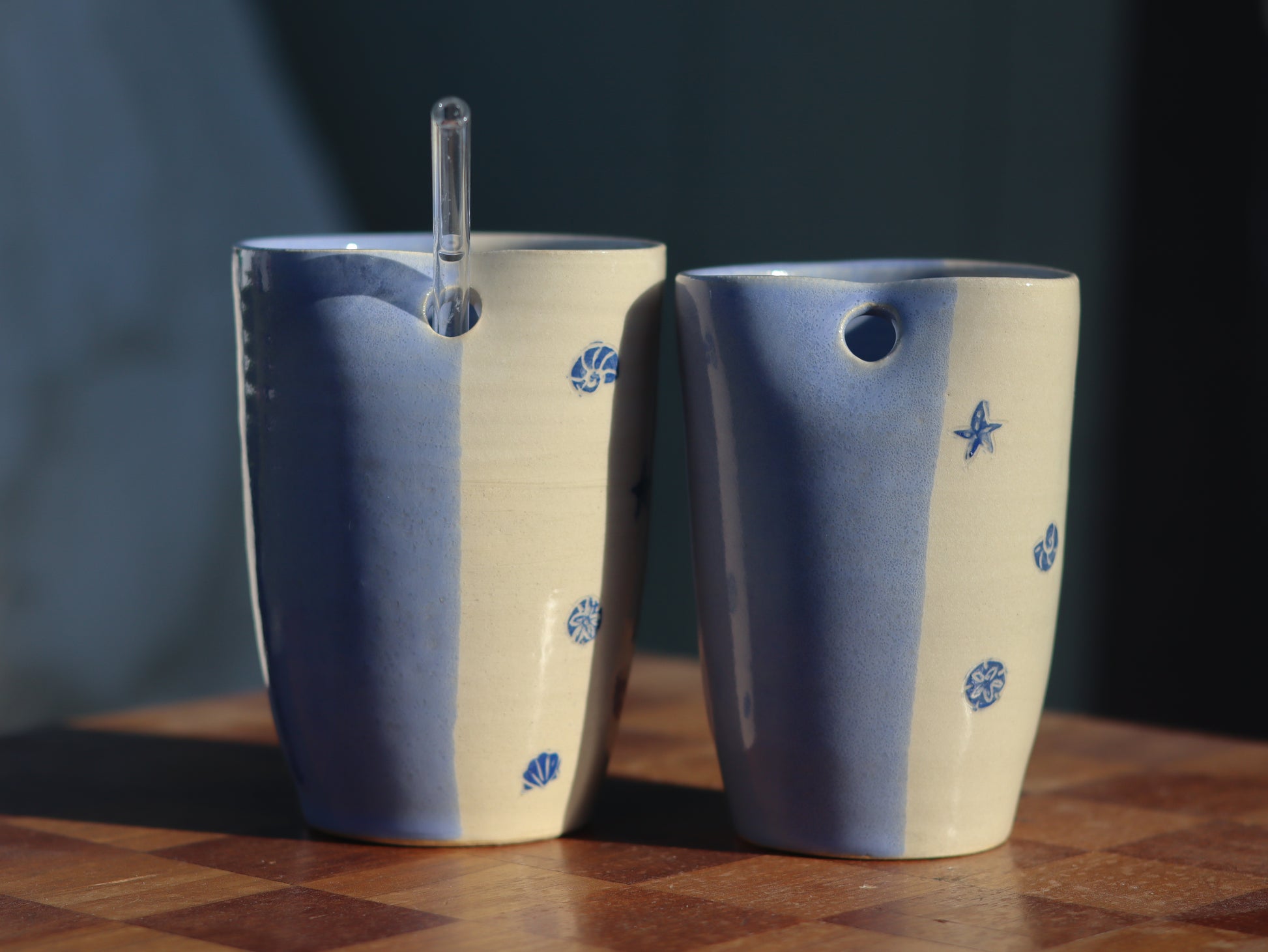 Two ceramic straw tumblers with a blue and cream color scheme, decorated with blue seashell and starfish motifs, displayed on a wooden surface.
