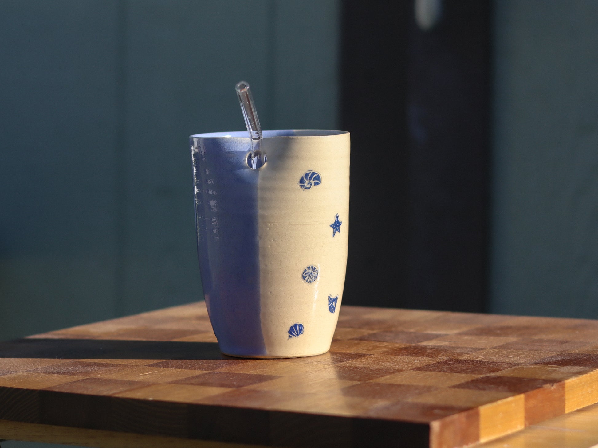 A handmade ceramic straw tumbler with a blue and cream color scheme, decorated with blue seashell and star motifs, sitting on a wooden cutting board with a glass straw inside.